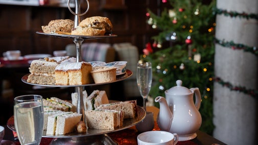 A tiered afternoon tea stand brimming with cakes, sandwiches and scones sits on a table with glasses of elderflower bubbly, a teapot and a cup of tea. There is a decorated Christmas tree in the background as well as a pillar wrapped in a festive garland.